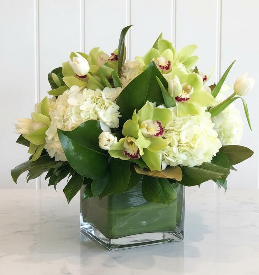 A floral arrangement of white hydrangeas and light green orchids with green leaves in a square glass vase, placed on a white tablecloth in front of a fireplace.