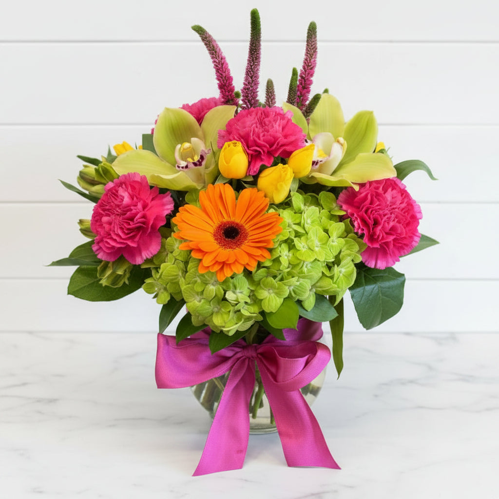 A vibrant floral arrangement featuring bright pink catnations, lime green orchids, a large orange gerbera daisy, yellow tulips, and green hydrangeas tied with a magenta ribbon, set against a rustic wooden background.