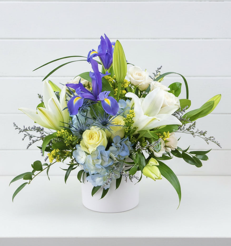 A mixed floral arrangement in a round white pot, featuring white lilies, yellow roses, blue hydrangeas, purple irises, and green foliage, displayed on a wooden surface against a light gray wall.