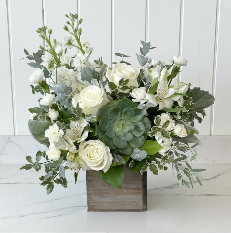 A floral arrangement featuring white roses, white alstroemeria, green succulents, and eucalyptus in a rustic square wooden box, displayed on a white pedestal against a light gray wall.