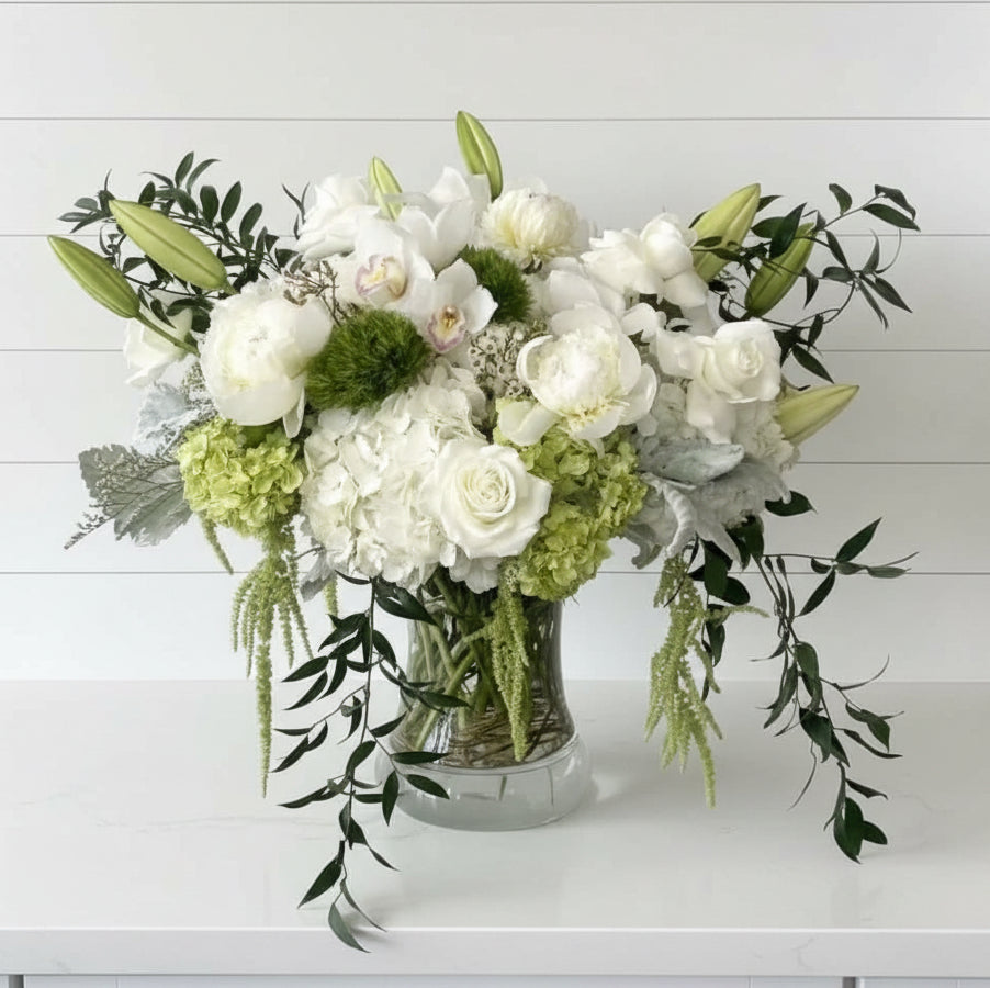 A large floral arrangement in a clear glass vase, featuring white roses, white orchids, green hydrangeas, and green foliage, displayed on a white pedestal against a light gray background.