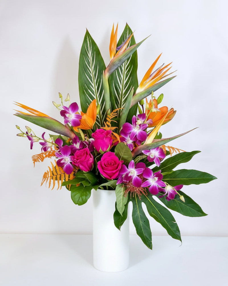 A vibrant tropical flower arrangement in a clear glass cylinder vase, featuring bright orange bird of paradise flowers, pink roses, green leaves, and orange pincushion protea on a white background.