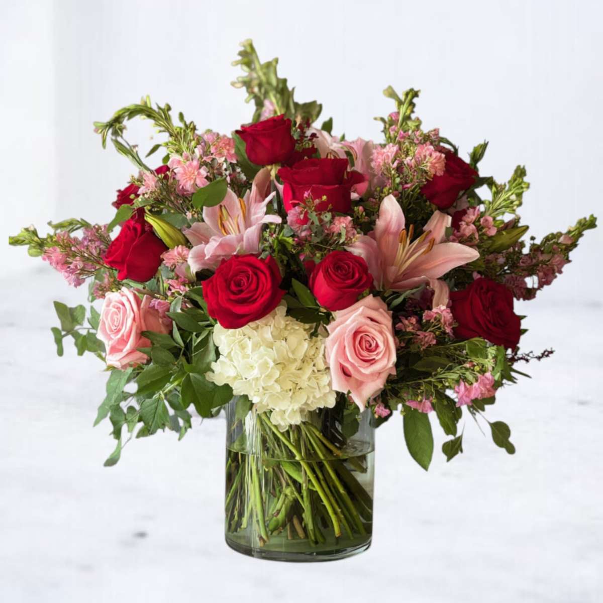 Bouquet of red roses, pink lilies, and white hydrangeas in a clear glass vase on a light gray background.