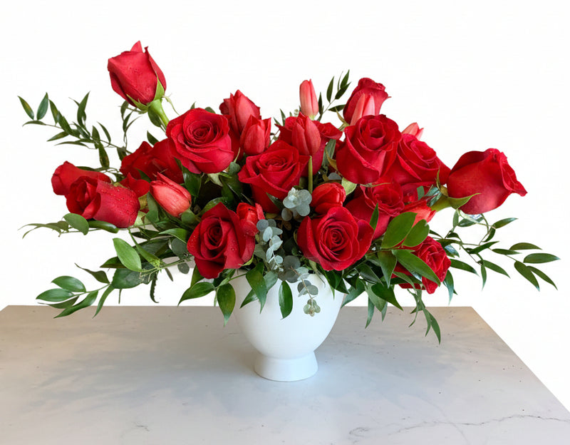 A vibrant red rose bouquet in a white ceramic vase, displayed on a white table next to a small candle with a card that reads 'esmé bouquets'.