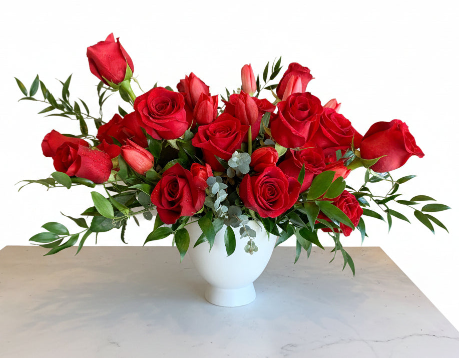 A vibrant red rose bouquet in a white ceramic vase, displayed on a white table next to a small candle with a card that reads 'esmé bouquets'.