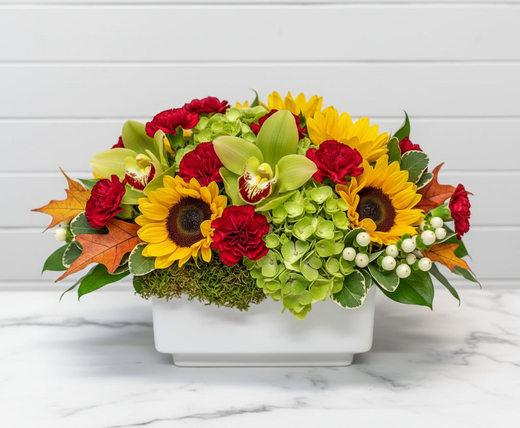 A vibrant autumn floral arrangement in a green glass vase, featuring yellow sunflowers, red roses, green orchids, orange anemones, and white berries, set against a white brick wall background.