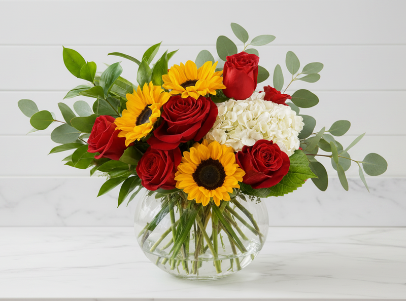 A vibrant mixed floral arrangement in a clear glass vase, featuring red roses, yellow sunflowers, white hydrangeas, and green foliage, displayed on a glass-topped side table against a neutral cream background.