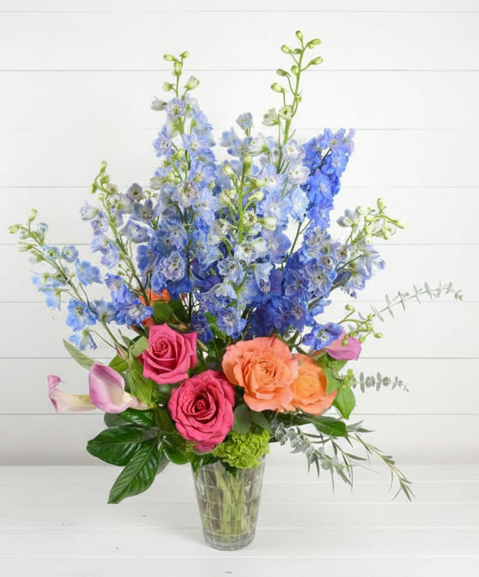 Bouquet of flowers including roses and delphiniums in a clear vase on a white background